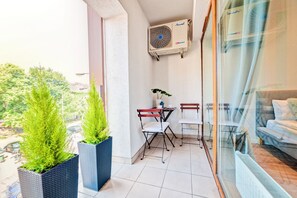 A balcony with potted plants, a table with chairs, a window with a view of greenery, and an air conditioning unit.