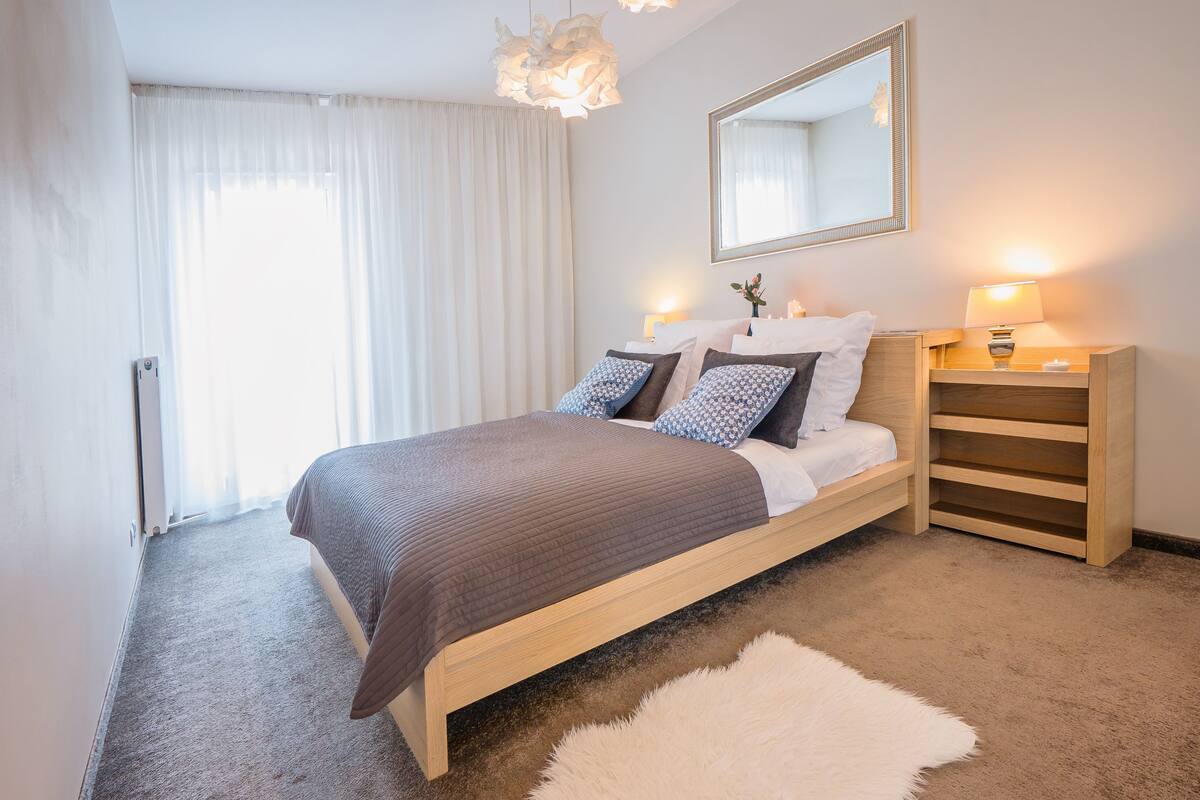 A bedroom with a bed, a bedside table with a lamp, a mirror, and a fluffy rug.