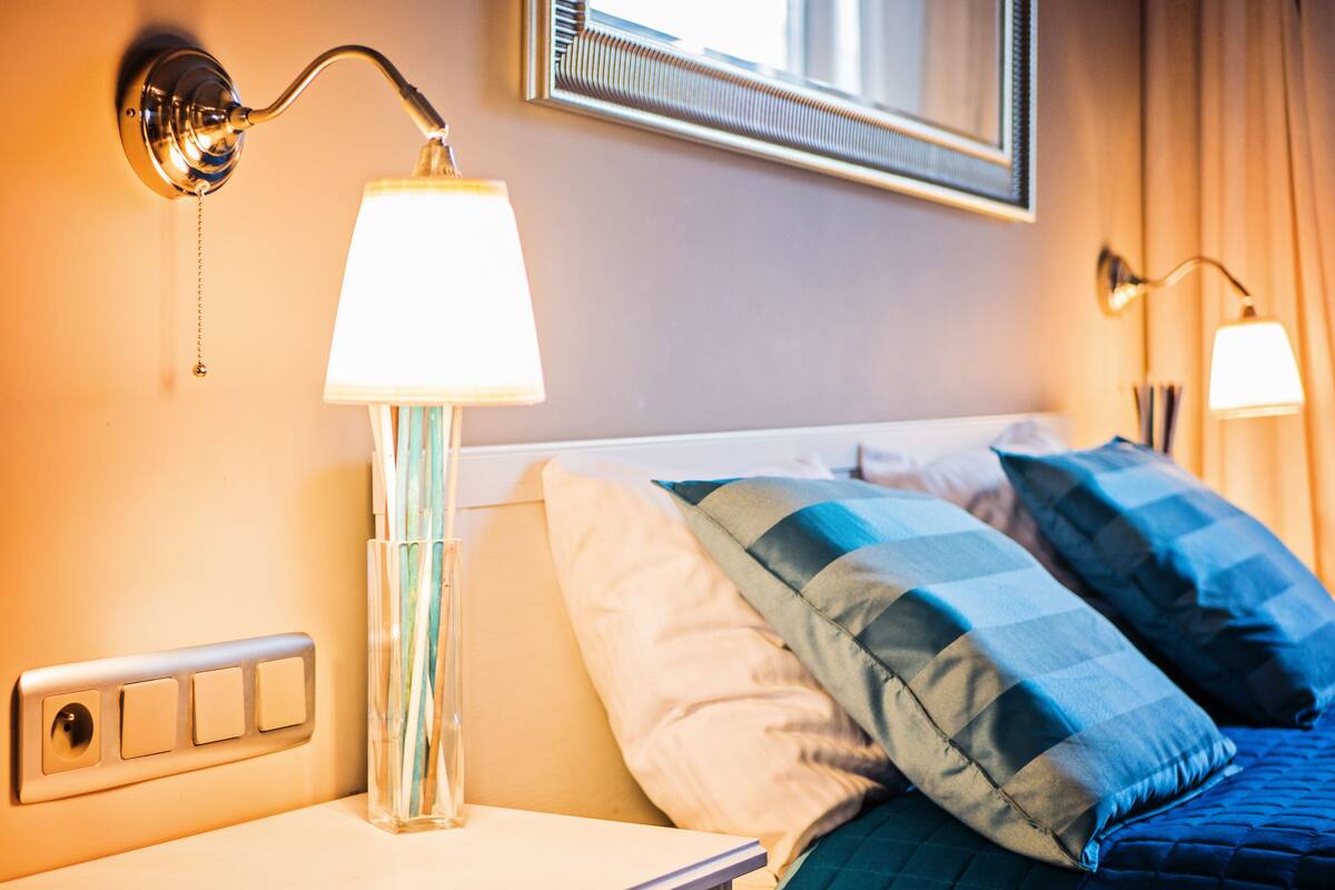 A hotel room with a bed, a nightstand, a lamp, and a wall-mounted light fixture.