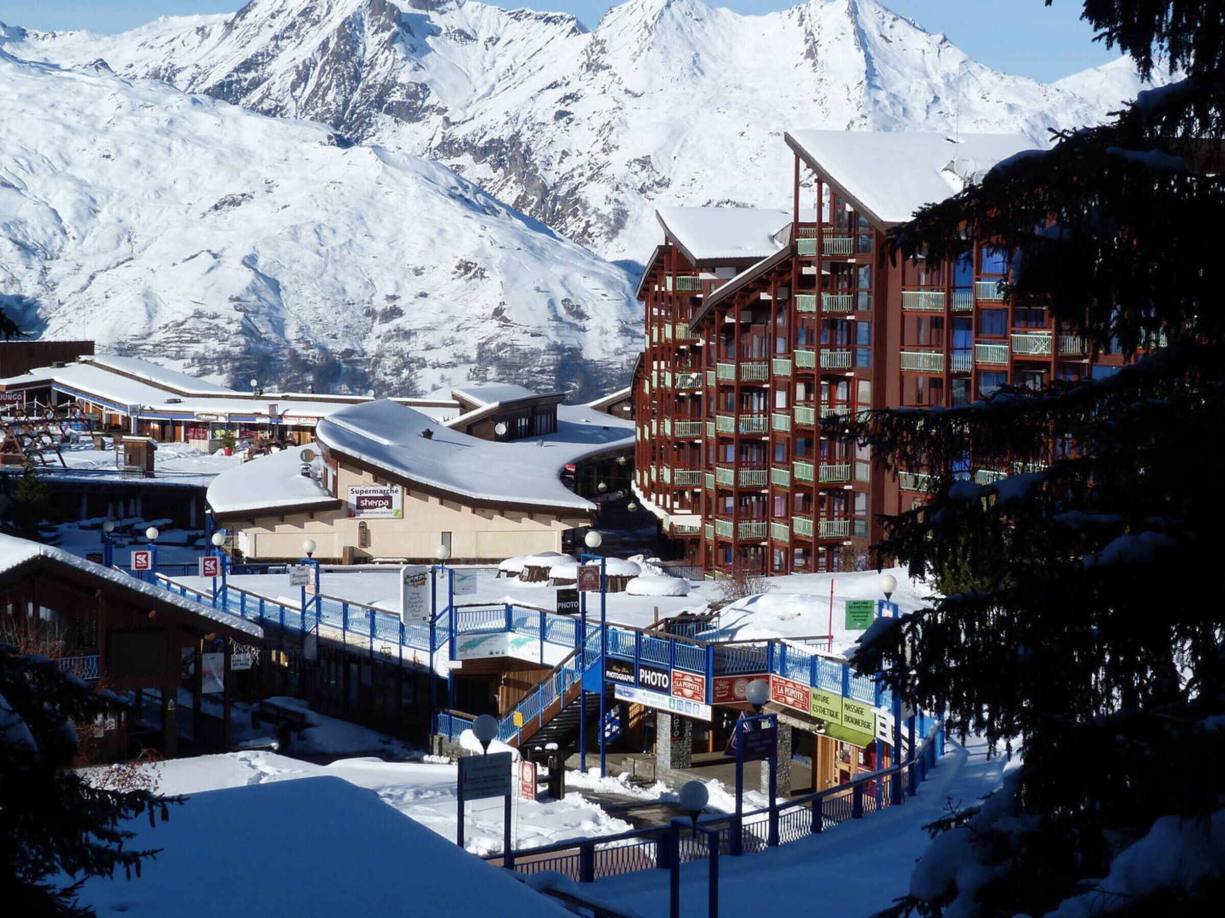 A snow-covered mountain town with a prominent red building with balconies, a ski lift, and a sign for 'Supermarché'.