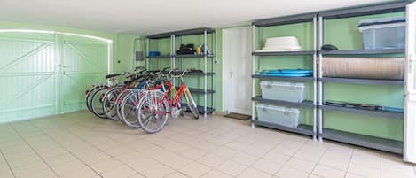 Quarto com porta verde, chão de azulejos e suporte para bicicletas com várias bicicletas. Há prateleiras com caixas organizadoras e uma porta branca ao fundo.
