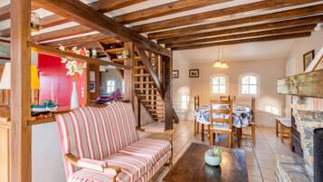 A cozy living room with a wooden staircase, a striped sofa, a dining table with chairs, and a stone fireplace.