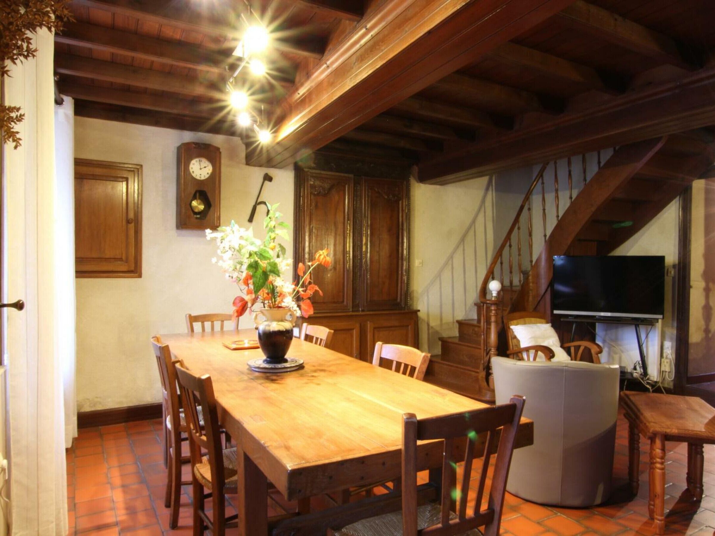 Un comedor con una mesa de madera, sillas, un jarrón con flores, un reloj y un televisor sobre un soporte.