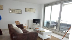 A living room with a brown sofa, white coffee table, and a TV. There are framed pictures on the wall and a large window. - 3-Bed Apt in Central Arcachon, Sea View, Parking, Pets Allowed, Wifi (Arcachon)
