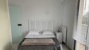 A small, modern bedroom with a bed, bedside table, and a window with blinds. - 3-Bed Apt in Central Arcachon, Sea View, Parking, Pets Allowed, Wifi (Arcachon)