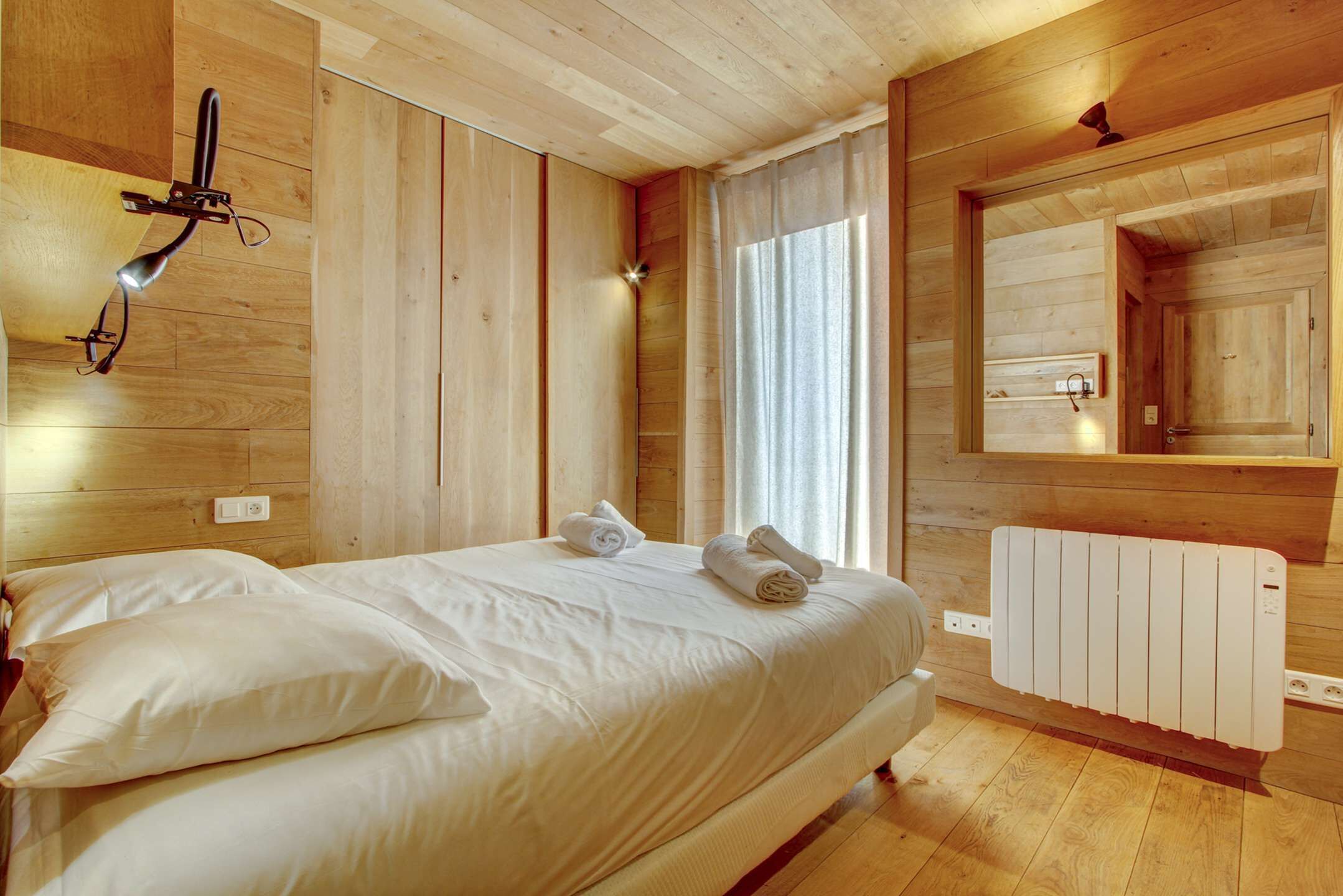 A wooden-paneled room with a bed, a wall-mounted light, a radiator, and a window with curtains.