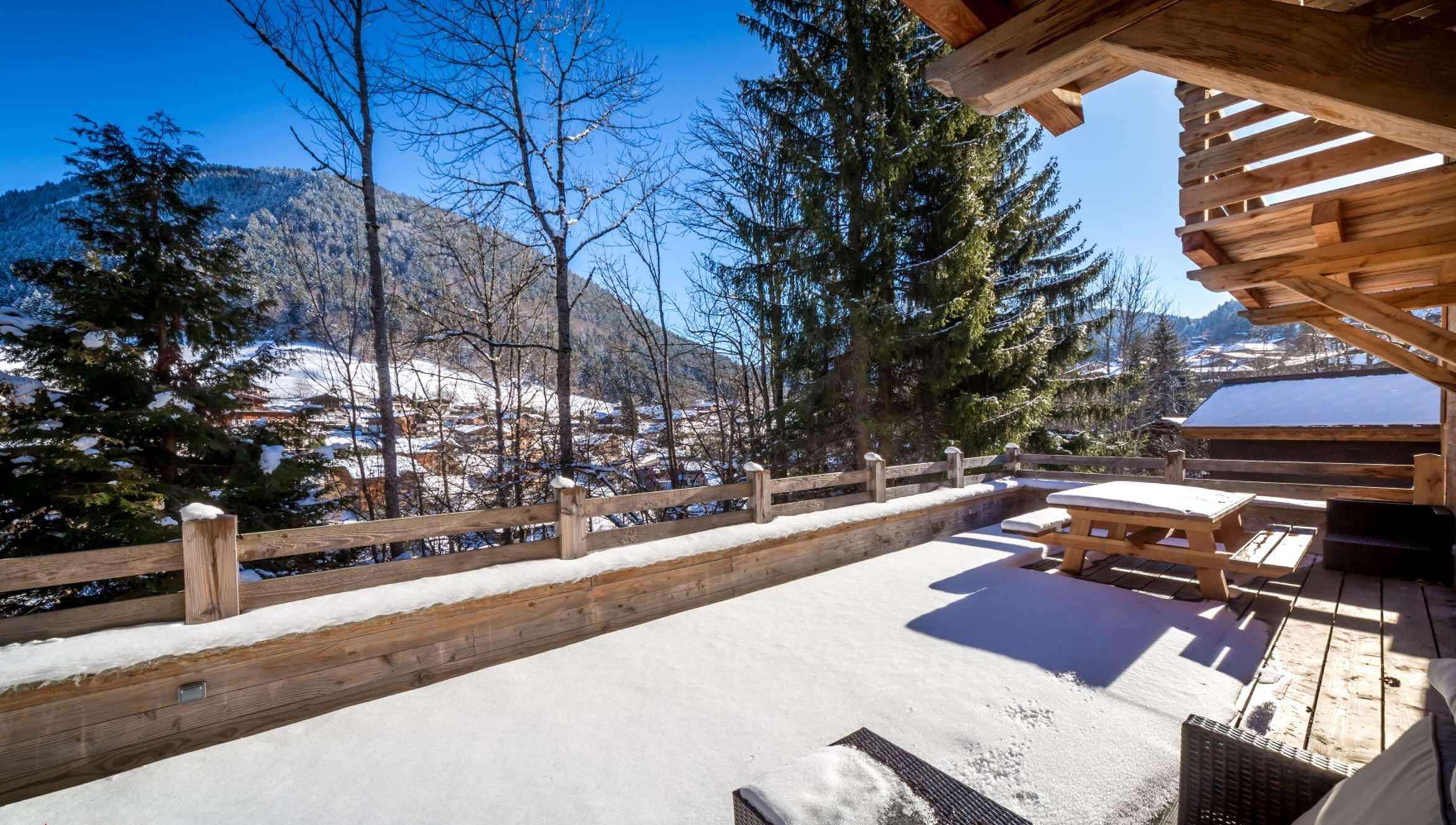 Une terrasse en bois recouverte de neige, une clôture en bois et une vue sur un village enneigé, avec des arbres et des montagnes en arrière-plan.