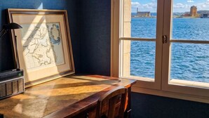 Una stanza con scrivania, sedia, cartina incorniciata e finestra vista su uno specchio d'acqua e edifici in lontananza.