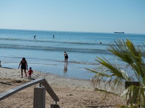 Une plage où l’on voit des gens se promener et nager, une clôture en bois et un navire au loin.