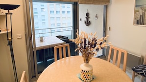 A dining area with a round table, chairs, a vase with dried flowers, a window with a view of buildings, and a floor lamp. - Sunny Apartment Near Beach, WiFi, Garage, Balcony (Les Sables-d'Olonne)