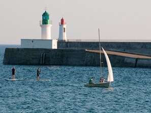 Un paysage côtier avec un phare, une jetée et des personnes pratiquant des activités nautiques.