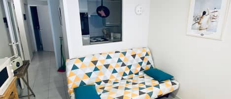 A living room with a sofa, a coffee table, a framed picture, and a kitchen area visible through an open door.