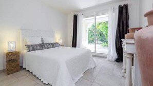 A bedroom with a large bed, a nightstand with a lamp, a dresser, and a view of greenery outside.