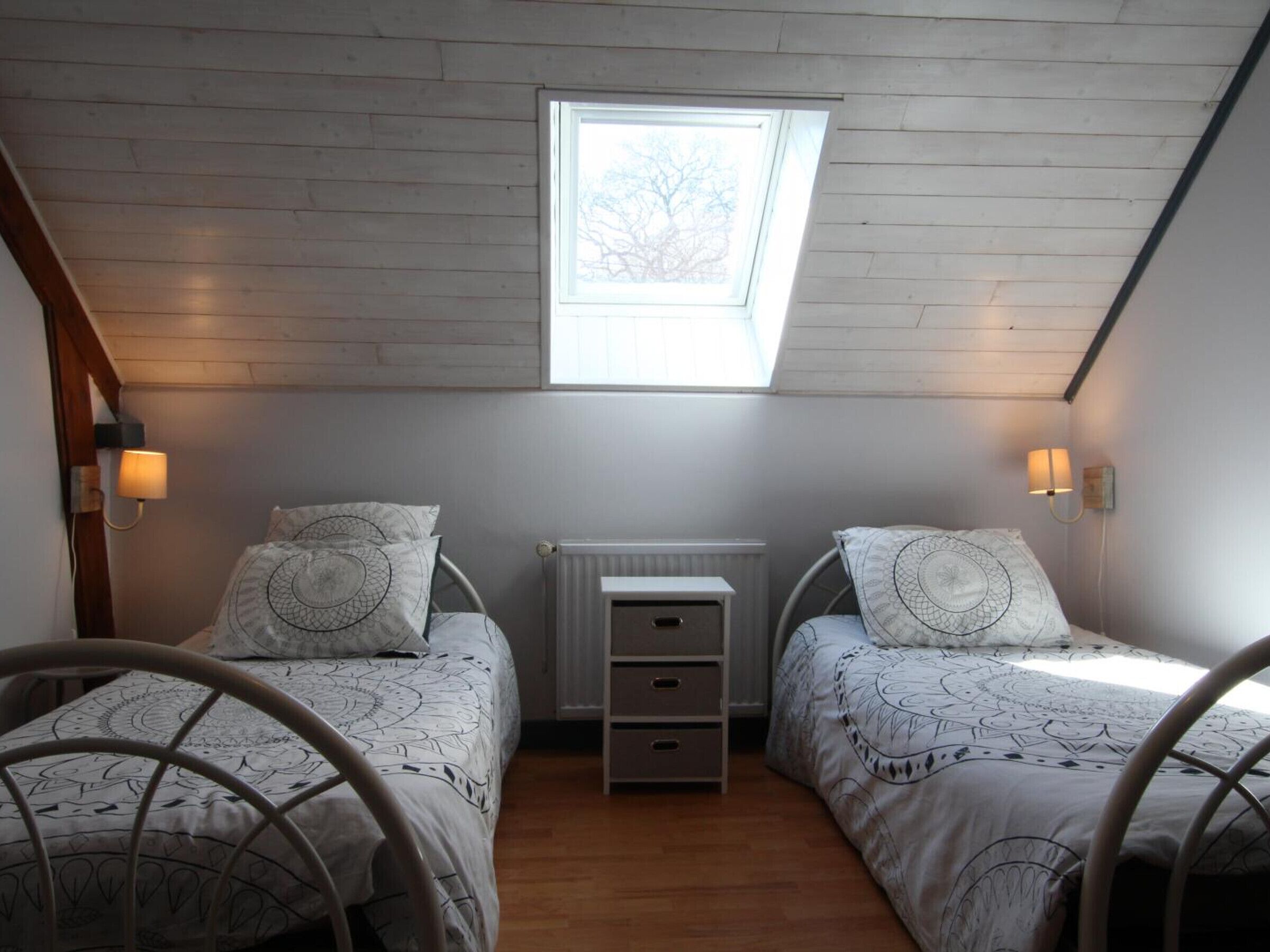 Une petite chambre confortable avec deux lits simples, une table de chevet, un radiateur et une lucarne.