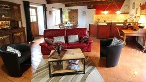 Una sala de estar acogedora con un sofá rojo, sillones negros y una mesa de centro de madera. Se ve una cocina con armarios rojos y una lámpara colgante al fondo.