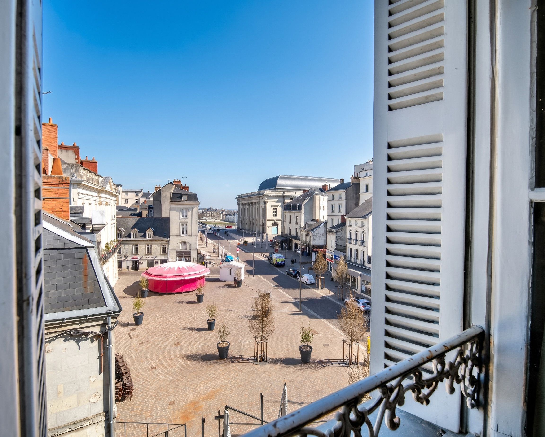 Une vue depuis un balcon donnant sur une rue de la ville, avec des immeubles, un parasol rouge et un ciel dégagé.