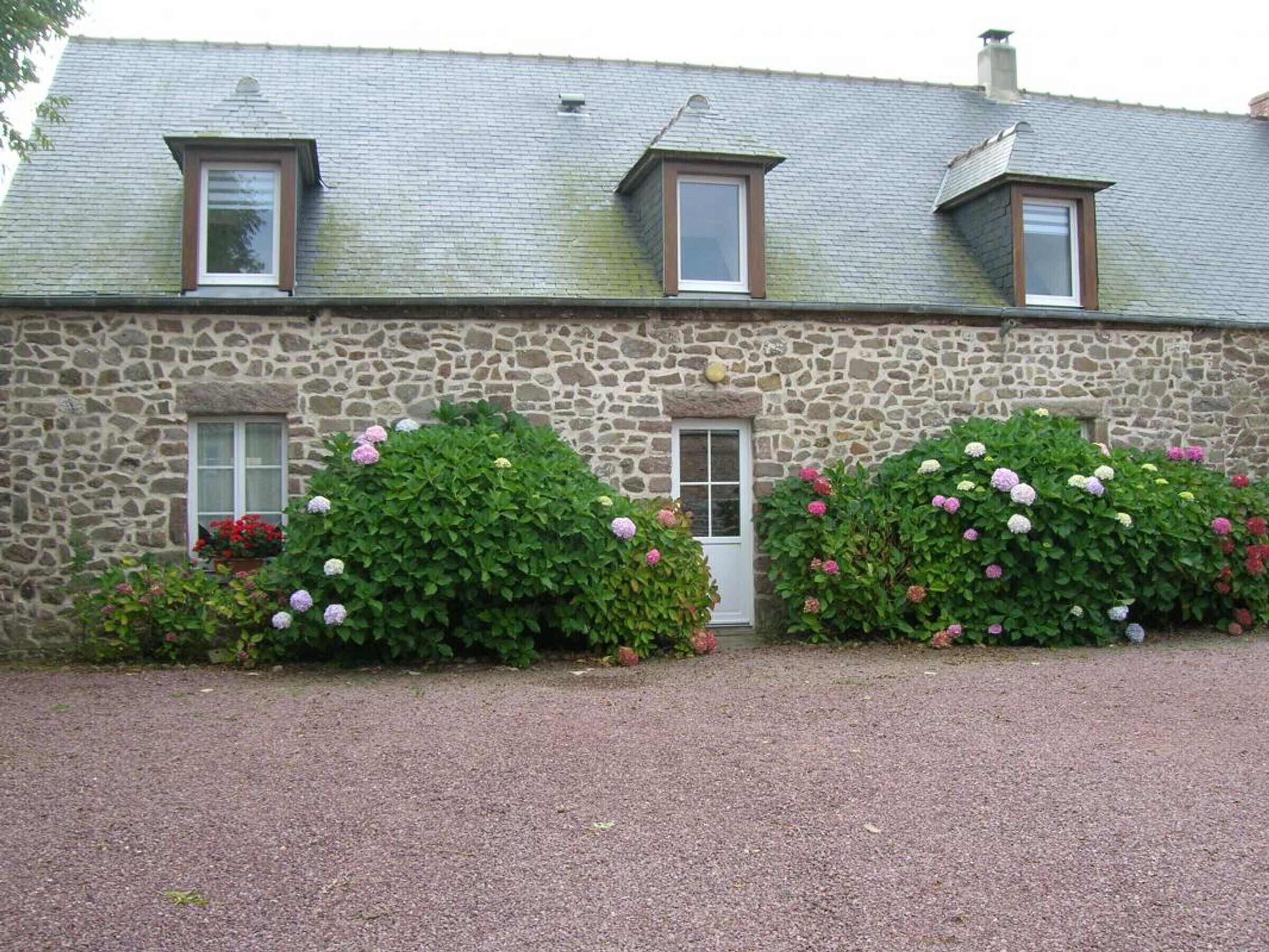 Un bâtiment en pierre avec une allée de gravier, deux fenêtres avec volets et une porte.
