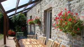 Une table et des chaises en bois sont disposées sous une pergola, avec un mur de pierres en arrière-plan, et des paniers de fleurs suspendus ajoutent une touche décorative.