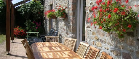 A wooden table and chairs set up under a pergola with a stone wall backdrop, featuring hanging flower baskets.