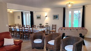 Un salon chaleureux avec un canapé rouge, des fauteuils en osier, une table de baby-foot et un coin repas comprenant une table et des chaises.