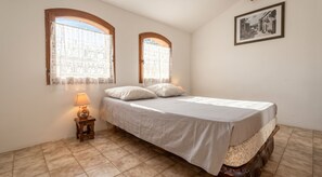 Un dormitorio con cama, dos ventanas con cortinas, una lámpara de noche y un cuadro en la pared.