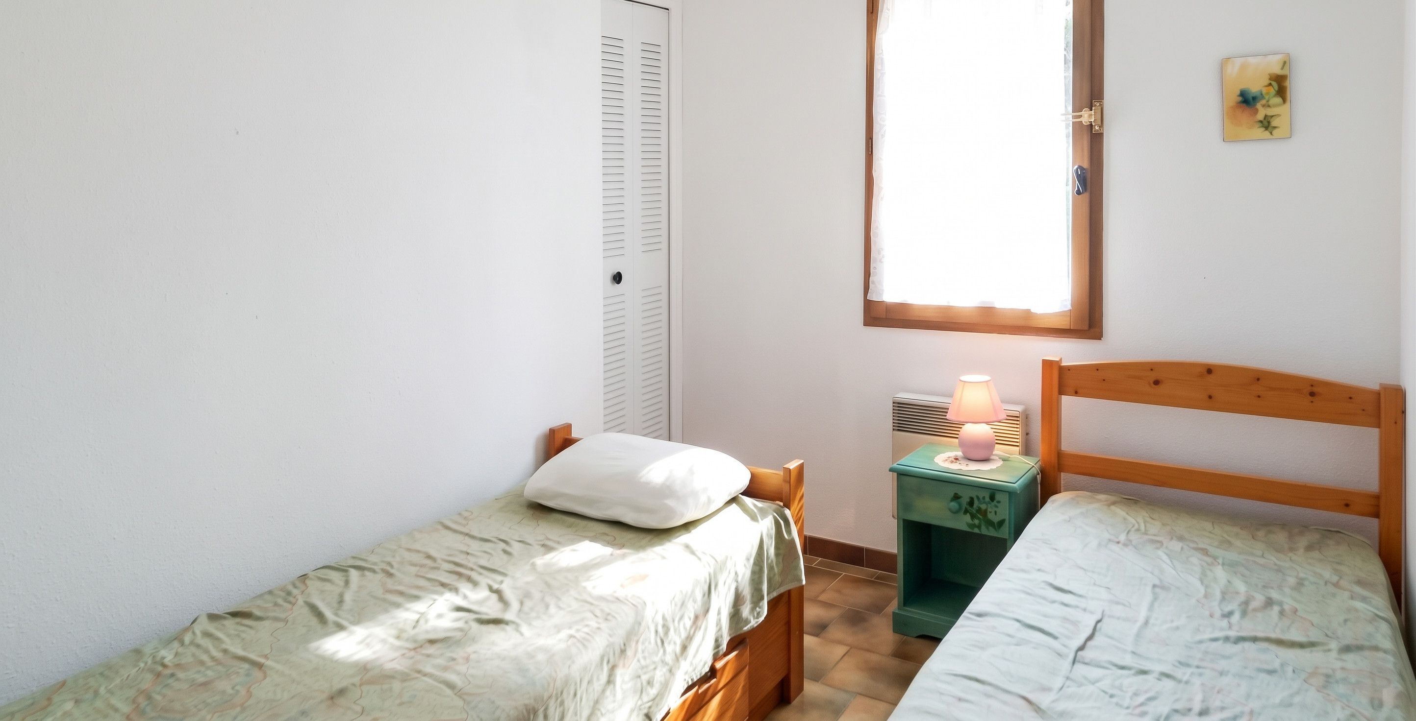Een kleine slaapkamer met twee eenpersoonsbedden, een nachtkastje met lamp, een kledingkast en een raam met uitzicht.