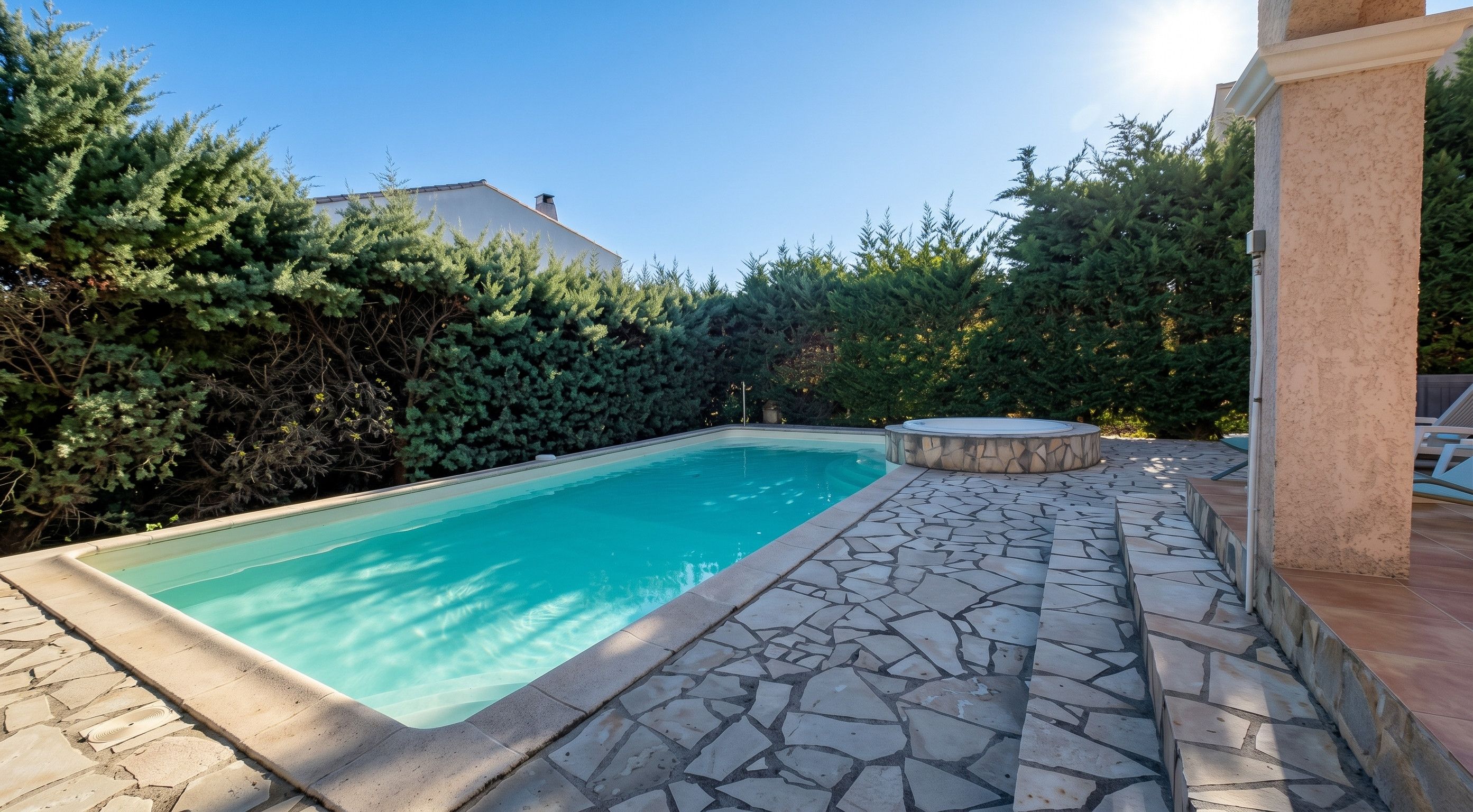 Une piscine aux carreaux de pierre et un jacuzzi en pierre, entourés d’une végétation luxuriante et sous un ciel dégagé.
