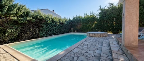 A swimming pool with stone tiles and a stone hot tub, surrounded by dense greenery and a clear sky.