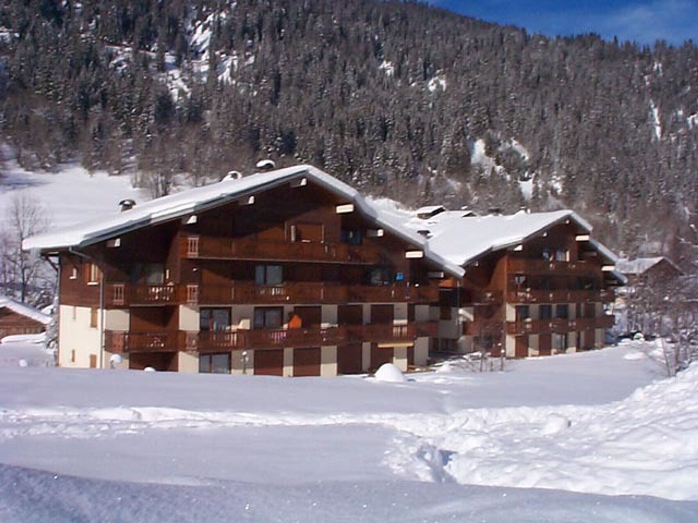 A wooden lodge with multiple balconies and a snow-covered roof, surrounded by a forested mountain backdrop.