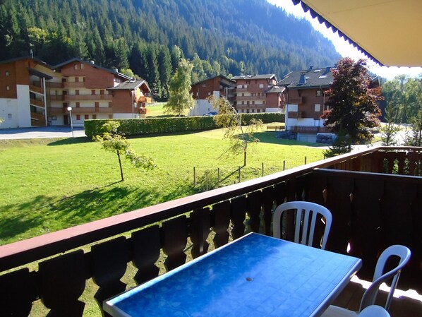 A balcony with a table and chairs, overlooking a green lawn and buildings in a mountainous area.