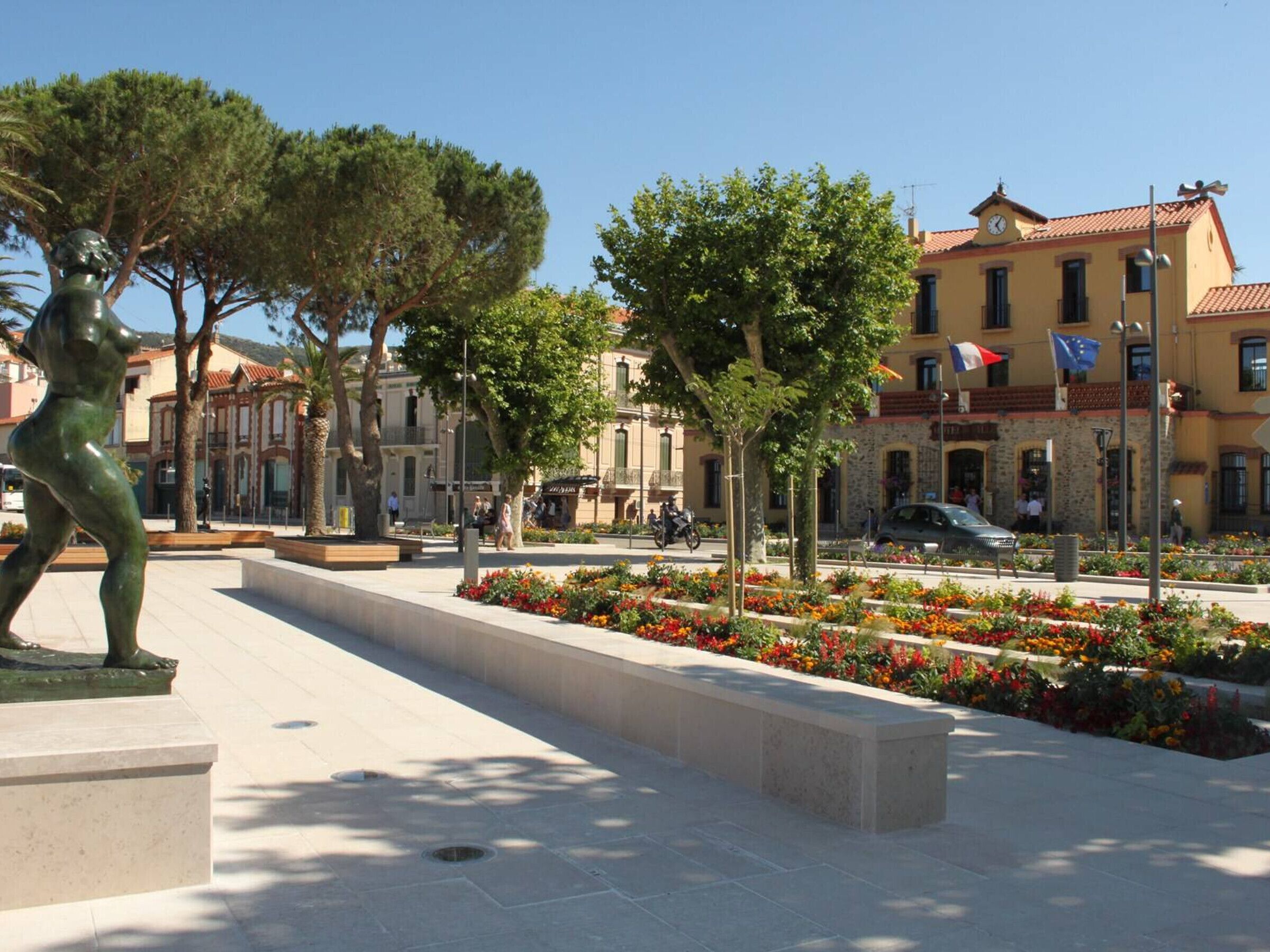 A public square with a statue, trees, and flower beds.