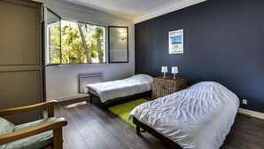A bedroom with two single beds, a window with a view of trees, a small table with two lamps, and a chair.