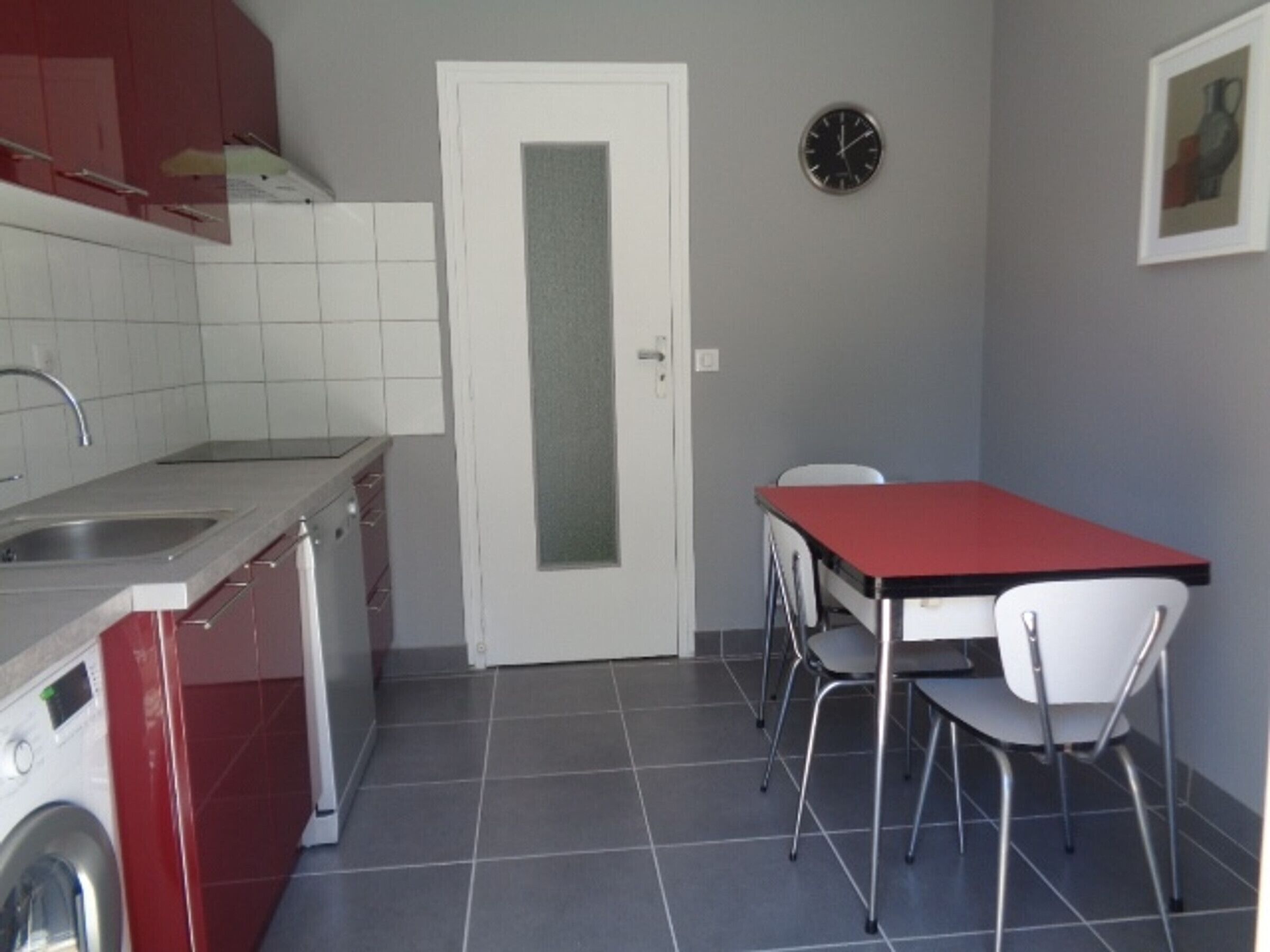 Ett kompakt kök med tvättmaskin, diskho, ett matbord med stolar, en klocka och en dörr.