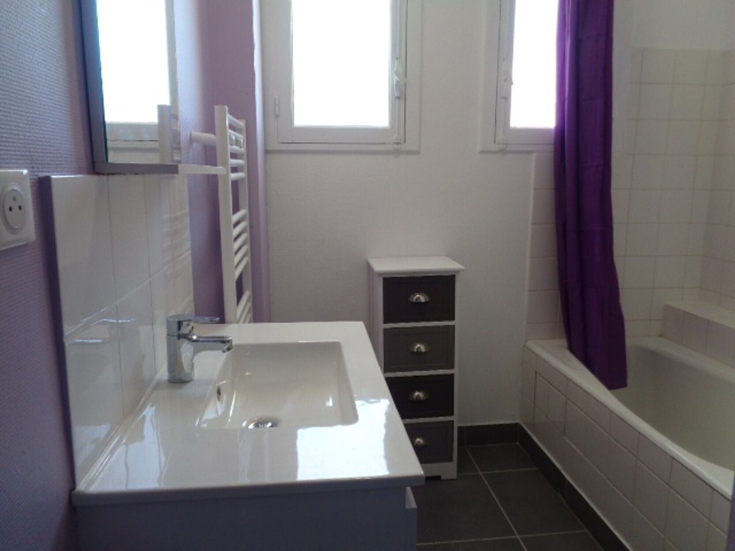 Een badkamer met een witte wastafel, een spiegel, een handdoekrek, een kast met lades en een bad.