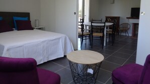 A bedroom with a bed, a small table, and purple chairs. There is a dining area with a table and chairs in the background.