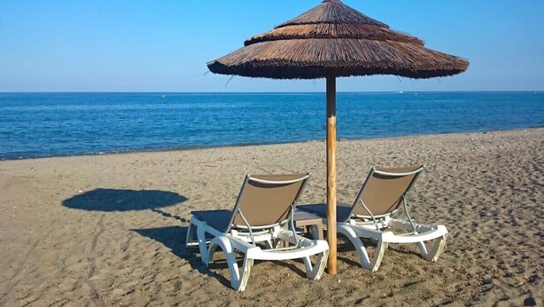 Two beach chairs under a thatched umbrella on a sandy beach with the ocean in the background. - Villa Jardins de la Mer (Poggio-Mezzana)