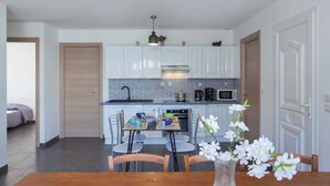 A modern kitchen with white cabinets, a tiled backsplash, and a dining area with a wooden table and chairs. - Villa Jardins de la Mer (Poggio-Mezzana)