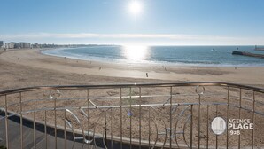 Uitzicht op het strand vanaf een balkon met een metalen hekwerk, een heldere lucht en de zon die op het water schittert.