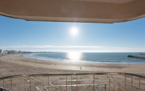 A beach view from a balcony with a metal railing, a clear sky, and the sun reflecting on the water.