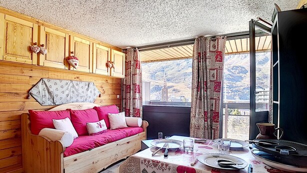 Un interior de cabaña acogedor, con un sofá de madera, una mesa de comedor para dos personas y un ventanal con vistas a la montaña.
