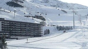 Un edificio moderno con techo plano y múltiples ventanas, rodeado por un paisaje nevado con pistas de esquí y un telesilla al fondo.