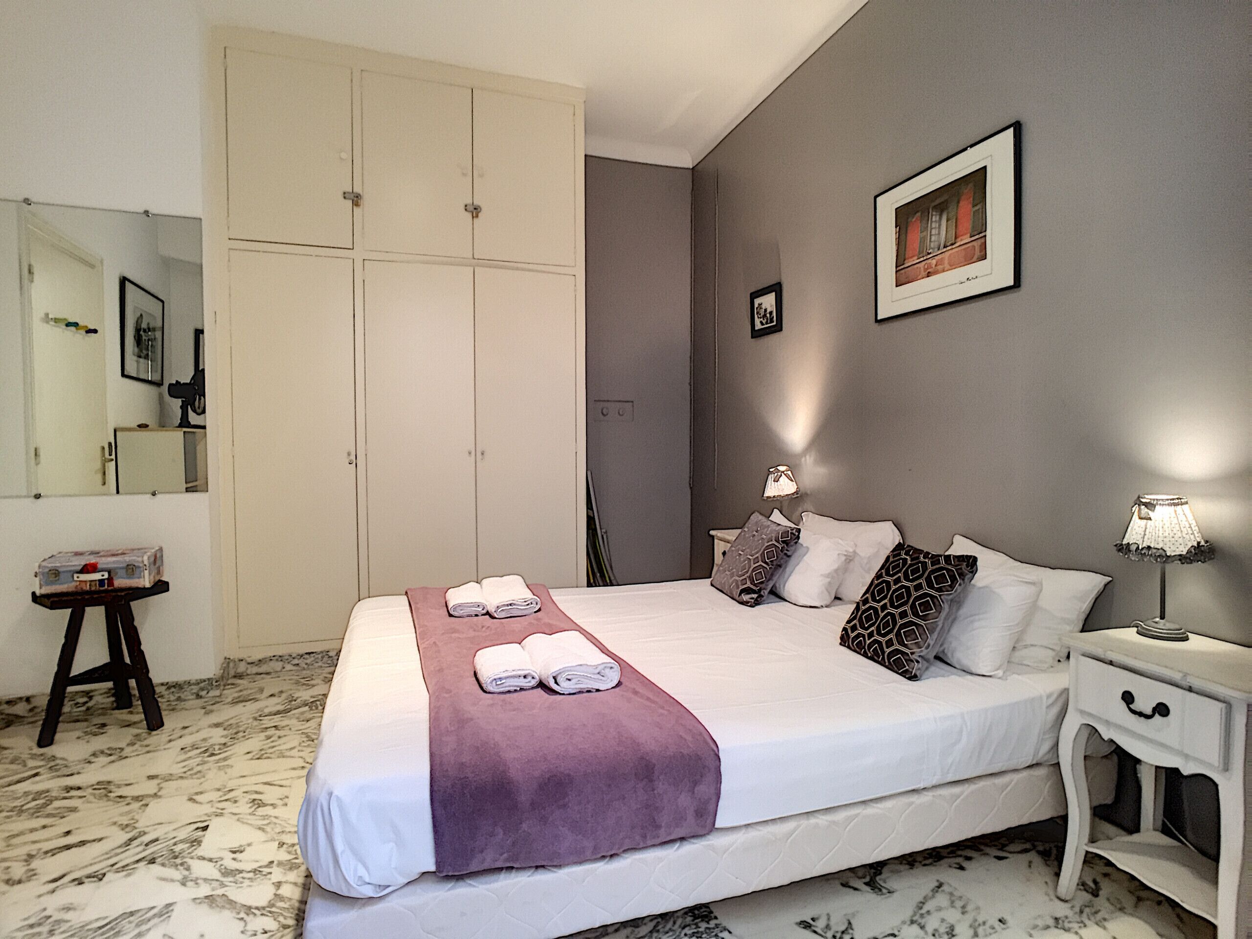 A neatly made bed with a purple blanket, a bedside table with a lamp, and a wardrobe in a hotel room.