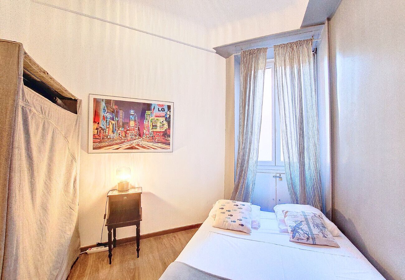 A hotel room with a bed, a small bedside table with a lamp, a window with curtains, and a cityscape painting on the wall.