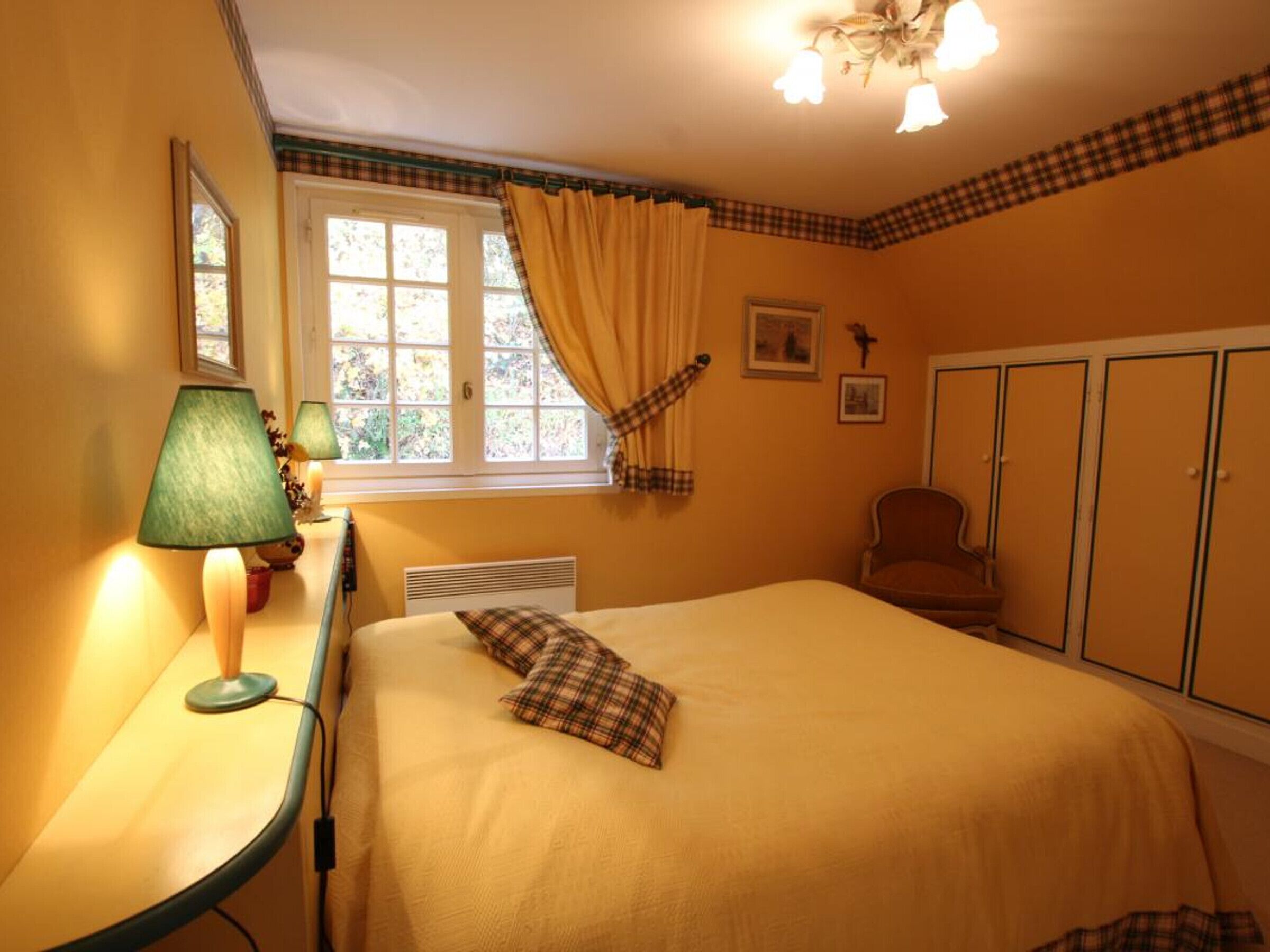 A bedroom with a bed, a desk with a lamp, a chair, a wardrobe, and a window with curtains.