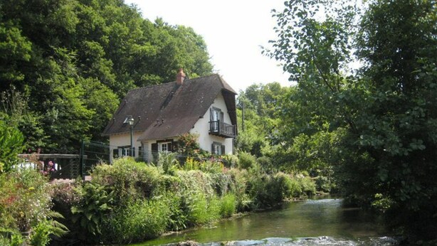 Een huis aan de rivieroever met rieten dak, omringd door weelderige begroeiing en een rustige rivier.