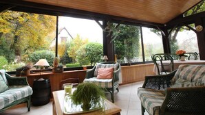 Een overdekt terras met rieten meubels, een houten tafel met een plant en uitzicht op een tuin en huis.