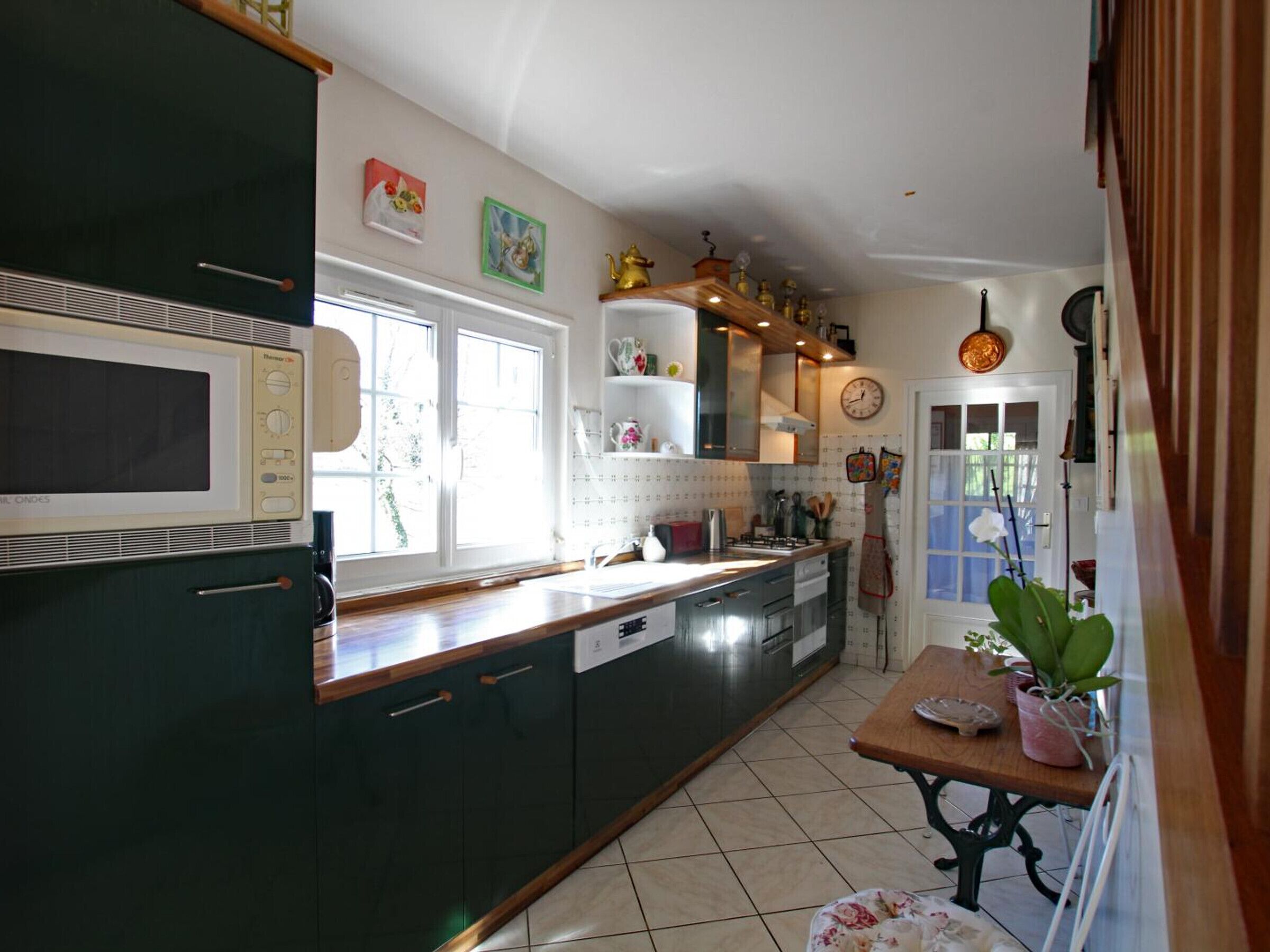 Een keuken met donkergroene kastjes, een witte magnetron, een houten aanrechtblad en een raam. Er is een spoelbak, een kookplaat en een klok aan de muur.