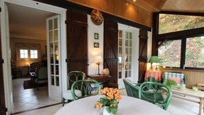 Een knus ingerichte kamer met een ronde tafel, groene stoelen en uitzicht op het groen buiten het grote raam.