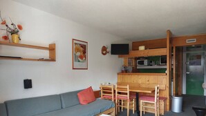 Un espacio habitable compacto con un sofá gris, una mesa de comedor de madera y sillas, una pequeña cocina con microondas, y una puerta verde.