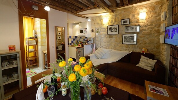 Una sala de estar acogedora con pared de piedra, techo de madera y un sofá. Hay una mesa de comedor con un florero y un televisor en la pared.